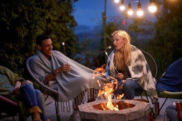 Young friends sitting outdoors on a summer night by the fire, roasting marshmallows and having fun together.