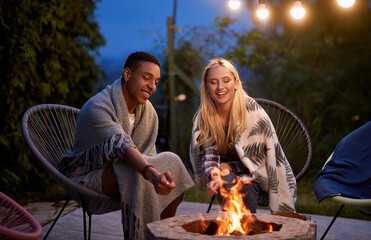 multiracial adult young couple, roasting marshmallow, smiling, having a fun, sitting by the fire covered with blankets