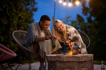 multiracial adult young couple, sitting by the fire, roasting marshmallow, having a fun