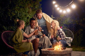 Young friends sitting outdoors by the fire on a cold summer night roasting marshmallows and toasting with beer.
