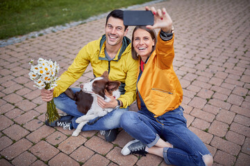 beautiful young female in casual outfit with ripped jeans on her knee taking selfie of herself and a boyfriend sitting with legs crossed and a dog in park