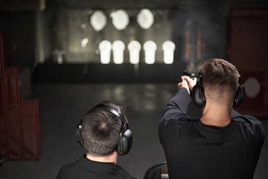 Two Caucasian men practicing shooting at indoor firing range, both wearing protective ear muffs, standing side by side with one aiming handgun toward paper targets in distance