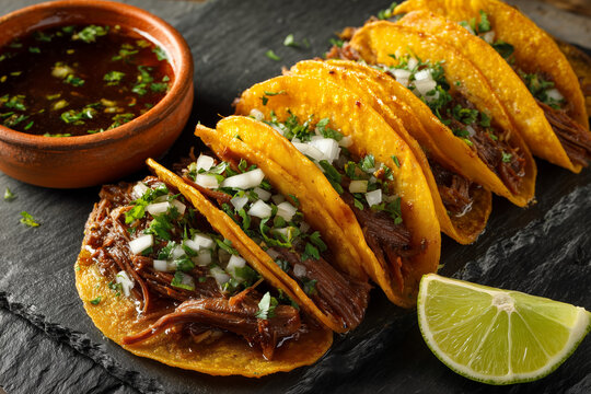 Juicy beef Birria tacos served with bowl Consomm&eacute; soup sauce, lime on dark black slate platter close up detail.