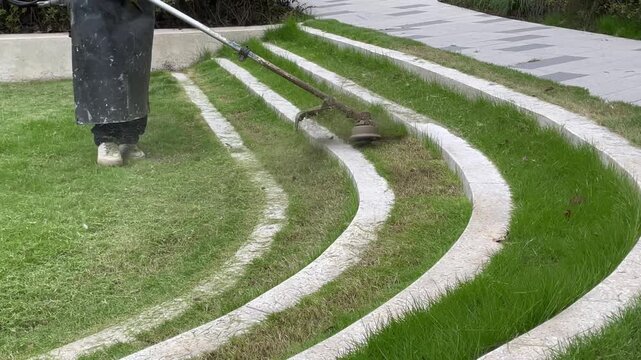 Worker trimming grass edges along stone steps and pavement with a string trimmer. Lawn care maintenance highlighting precise landscaping and clean borders between grass and concrete.