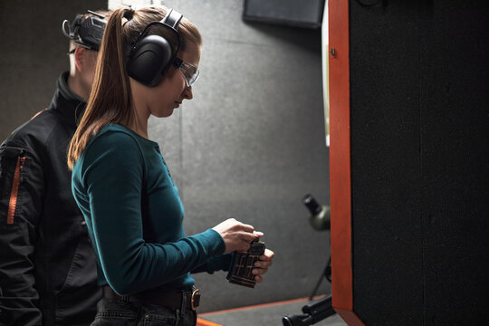 Young adult Caucasian woman loading handgun magazine at indoor shooting range, wearing protective earmuffs and safety glasses, standing next to young adult Caucasian man instructor
