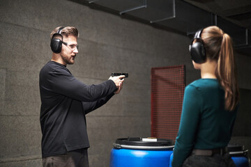 Caucasian young adult man instructing Caucasian young adult woman at indoor shooting range, both...