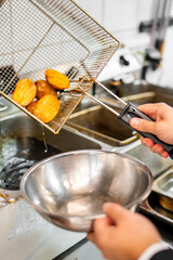 Golden-brown potato halves in a metal basket being drained after deep-frying in a commercial kitchen. Stainless steel bowl held below for transfer. Professional cooking process.