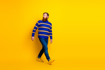 Stylish red-haired man walking confidently against a vibrant yellow backdrop wearing a striped sweater and jeans