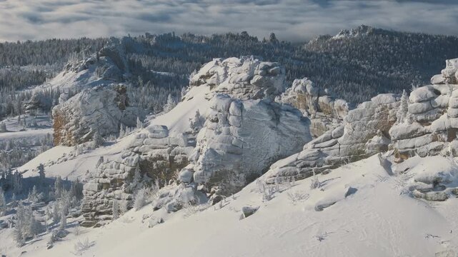 Aerial view of granite tower and snow-laden taiga at Sheregesh, the drone sliding along cliffs to reveal layered ledges and warm stone under rime. Bright morning light creates a cinematic alpine mood