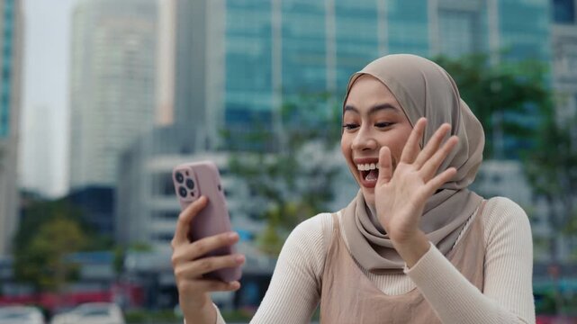 Portrait of happy Muslim woman uses video chat on smartphone stands against skyscrapers. Young Malaysian female answers video call waves Hello on city street. Online communication