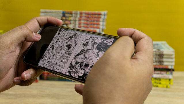 Kalimantan Selatan, Indonesia - 09 11 2025 : Digital screen showing Dandadan manga being read with pile of Japanese comics behind, representing online reading lifestyle.