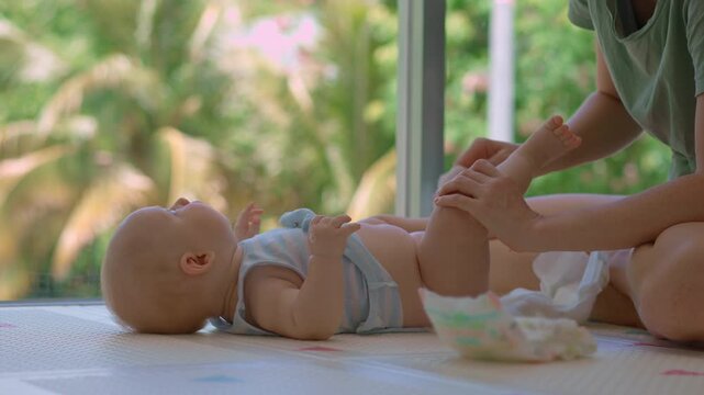 Young mother changes the diaper of her 3-month-old son by a large window with tropical greenery outside, creating a peaceful and loving baby care moment in a bright family home. Slowmotion video.