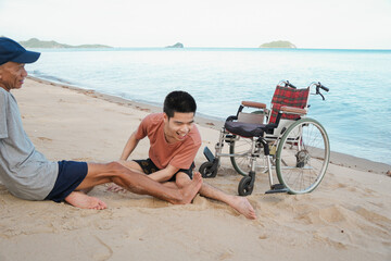 Asian young man sit on the beach near his wheelchair and his dad in vacation, Concept family...