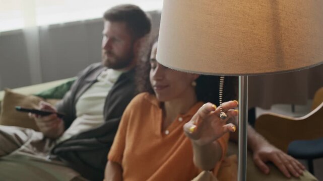 Relaxed couple watching TV together at home, with man holding remote control while woman turning on lamp with warm light, creating cozy atmosphere in living room