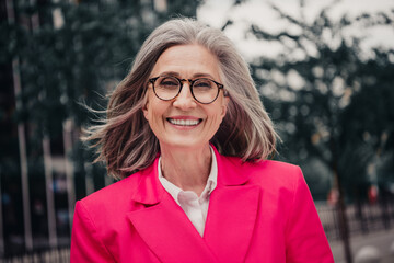 Confident senior woman with glasses and pink suit standing outdoors in urban setting on a sunny day