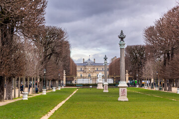 An alley of bare trees and ornamental columns leads to the Luxembourg Palace in the background. Unrecognizable people are walking on a cloudy day in the Luxembourg Gardens in Paris, France.