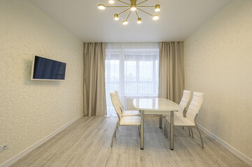 bright dining area features a white table with four chairs, light walls with a mounted TV, a window with curtains, and a modern chandelier