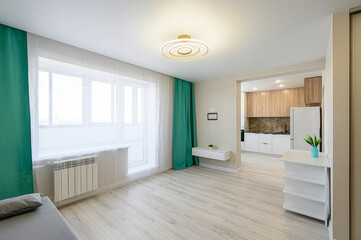 A modern, bright apartment interior with an open kitchen and living area, featuring light wood floors, white walls, and teal curtains