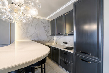 A modern kitchen with gray cabinets, marble backsplash, and a unique bubble chandelier. The kitchen features a curved counter with seating and integrated lighting.
