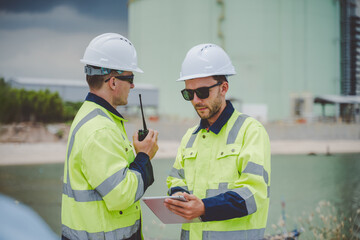 Two engineers using radio and tablet at coastal oil gas facility in safety jackets