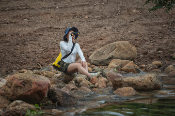 Young woman carrying a yellow backpack and camera is hiking through nature, carefully crossing a rocky stream in the forest. Outdoor adventure and eco-travel lifestyle concept.	