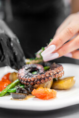 Chef garnishing grilled octopus with herbs using tweezers. Served with roasted potatoes, green beans, cherry tomatoes, and olives. Fine dining plating, close-up, professional kitchen.