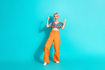 Cheerful young woman posing in vibrant orange pants and patterned top on a turquoise background exuding joyful energy