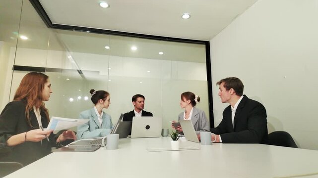 Boardroom Strategy Session: A focused team of professionals engage in a discussion around a conference table, laptops open, during a collaborative business meeting in a modern office setting.