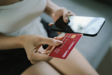 A young woman uses her smartphone and holds credit cards while sitting indoors, representing online shopping, finance, technology, and e-commerce.