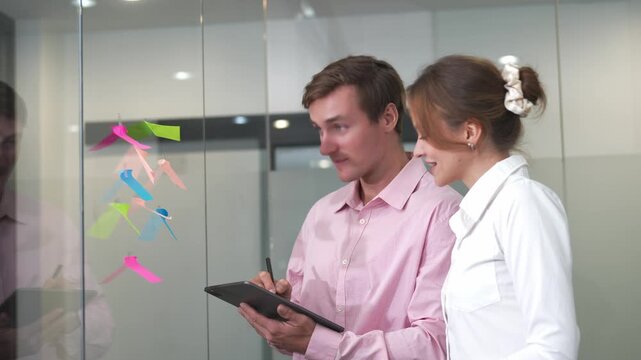 Collaborative Idea Generation: A man and a woman in a business setting brainstorm ideas using sticky notes and a tablet, fostering a dynamic exchange of thoughts and strategies for project success.