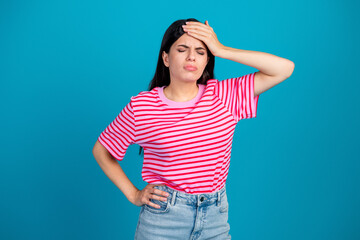 Young woman frowning while holding her head against a blue backdrop wearing pink striped shirt and denim shorts