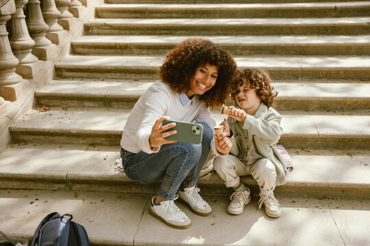 Mother and son holding ice cream and posing while sitting on a blanket on the stairs and looking at the phone she is holding