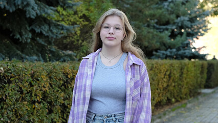 A young woman wearing glasses and a plaid shirt stands outdoors on a quiet path surrounded by...