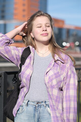 A blonde girl in casual attire tilts her face towards the sun, enjoying the warmth amidst a vibrant city setting. The image captures a balance of individuality and modern life.