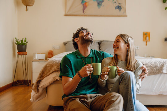 A man hugs a woman while they sit and lean on the bed while holding cups and laughing