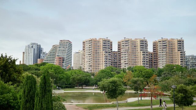Valencia, Spain - May 28, 2025: View of the Jardi del Turia showing a pond and sculpture framed by lush trees, with modern residential towers on the skyline under bright overcast light