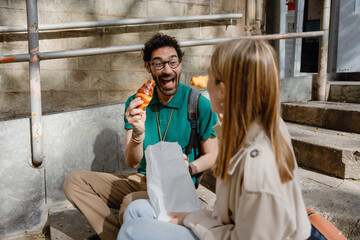 Obraz premium A man holds a croissant and laughs while a woman sits next to him on the stairs holding a package