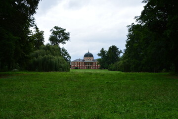 A grand and unique red chateau with a large dome sits in stately isolation at the far end of a vast green park lawn