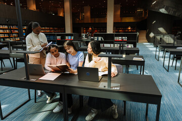 A group of three female students sit at desks and look at a book one of them is holding while a...