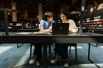 A female student is talking to a classmate who is looking at a laptop while they are sitting at a table