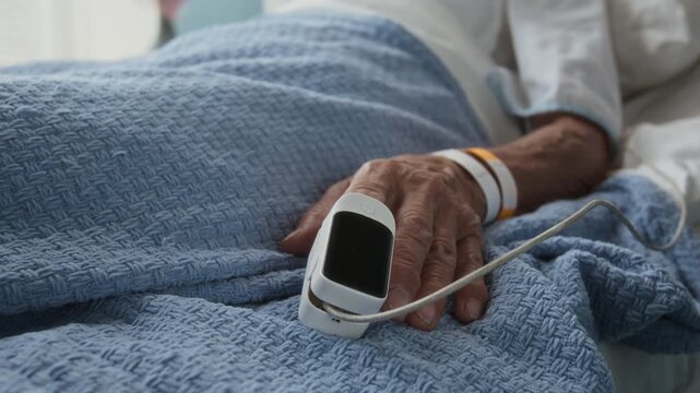 Close-up view of unrecognisable senior lady lying in hospital bed under blue blanket with pulse oximeter measuring oxygen saturation level of her blood