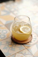 A cold lemon drink served in a round glass with ice cubes on a wooden coaster over a pastel patterned table, showing a fresh and summery beverage concept.