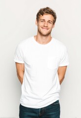 Obraz premium A young man stands confidently against a plain white background, smiling warmly at the viewer. He wears a simple white t-shirt with a pocket and dark wash jeans. His hair is tousled 