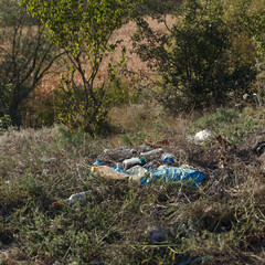 Littered area with plastic waste, glass and debris among dry grass and shrubs. Scene highlights environmental pollution