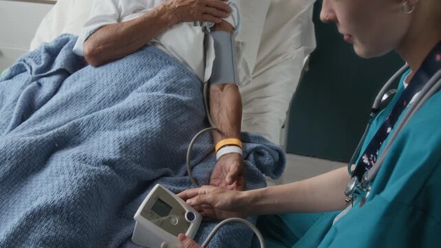 Tilt up view of young nurse using electronic sphygmomanometer to measure arterial blood pressure of senior female patient receiving treatment in hospital
