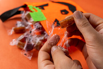 Hand taking and opening wrapped candies from orange felt jack-o'-lantern bag with an orange background.