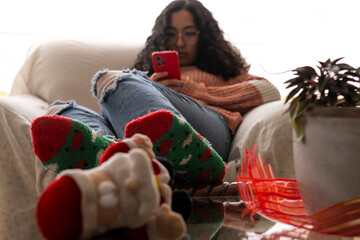 A young woman lounging on a sofa, wearing festive Christmas socks, browsing her phone, surrounded by a warm and cozy atmosphere 
