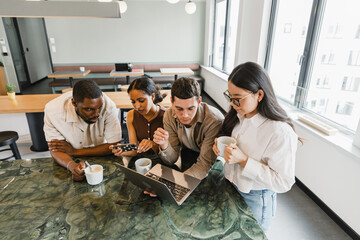 Obraz premium A group of four workers stand at a table while two of them hold cups, one of them holds a laptop, and one of them holds a phone
