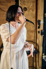 Obraz premium Young asian woman in white dress passionately sings in wooden studio. Warm lighting accentuates cozy atmosphere, while her focused expression highlights dedication and talent