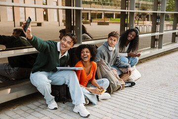A group of four students pose and smile while looking at a phone held by one of them while sitting near a building